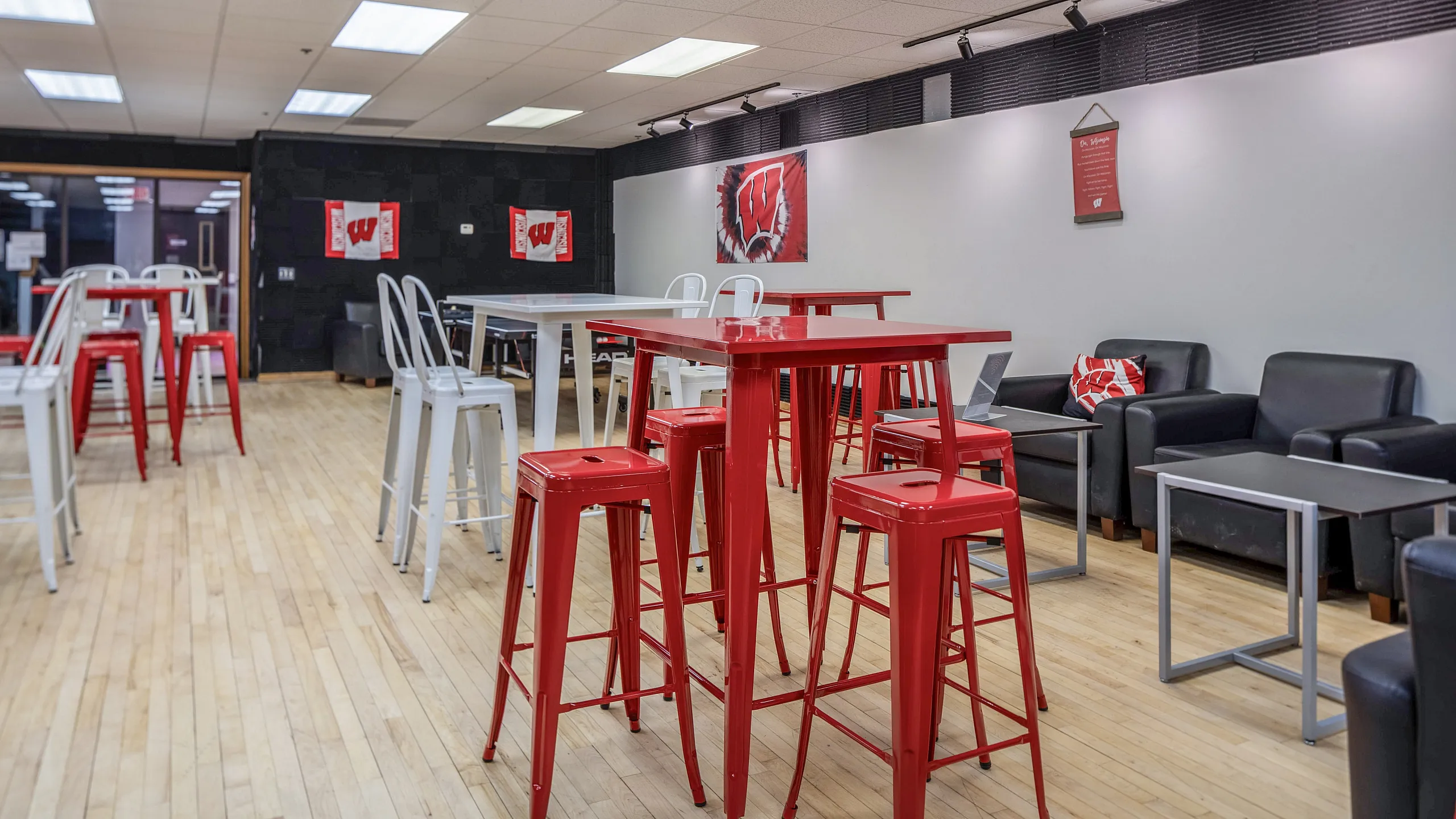 Stylish lounge area with modern red and white high tables and stools, featuring sleek black sofas and wooden floors.