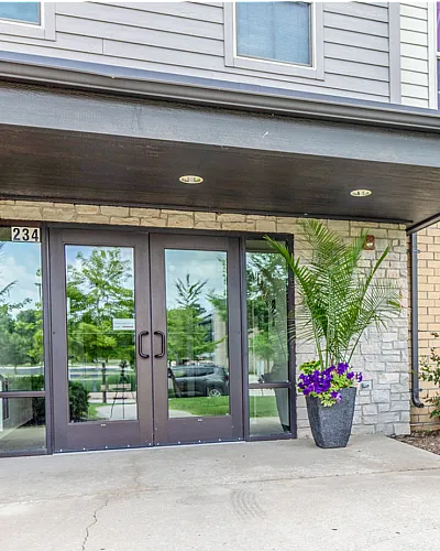 Inviting apartment entrance with elegant double glass doors, surrounded by vibrant landscaping and stylish planters, offering a warm welcome.