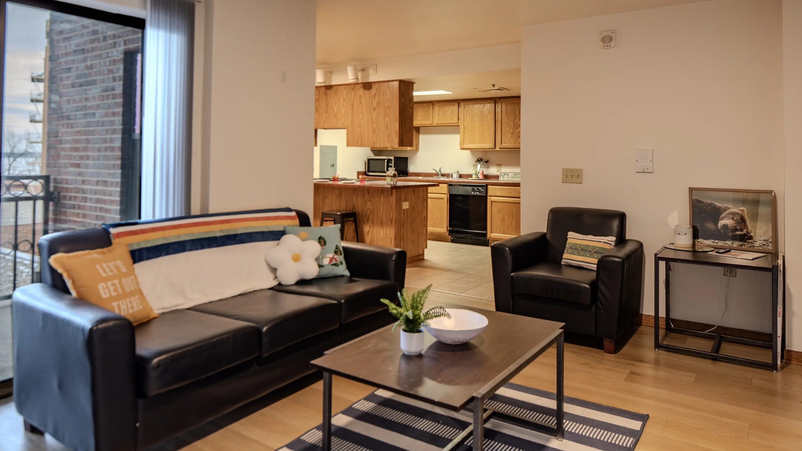 Charming living room with modern leather sofas and a cozy open kitchen, featuring wooden cabinets and a sunny balcony view.