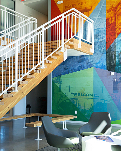Modern lobby with vibrant mural and sleek wooden staircase, complemented by stylish grey chairs and a round table, creating a welcoming atmosphere.