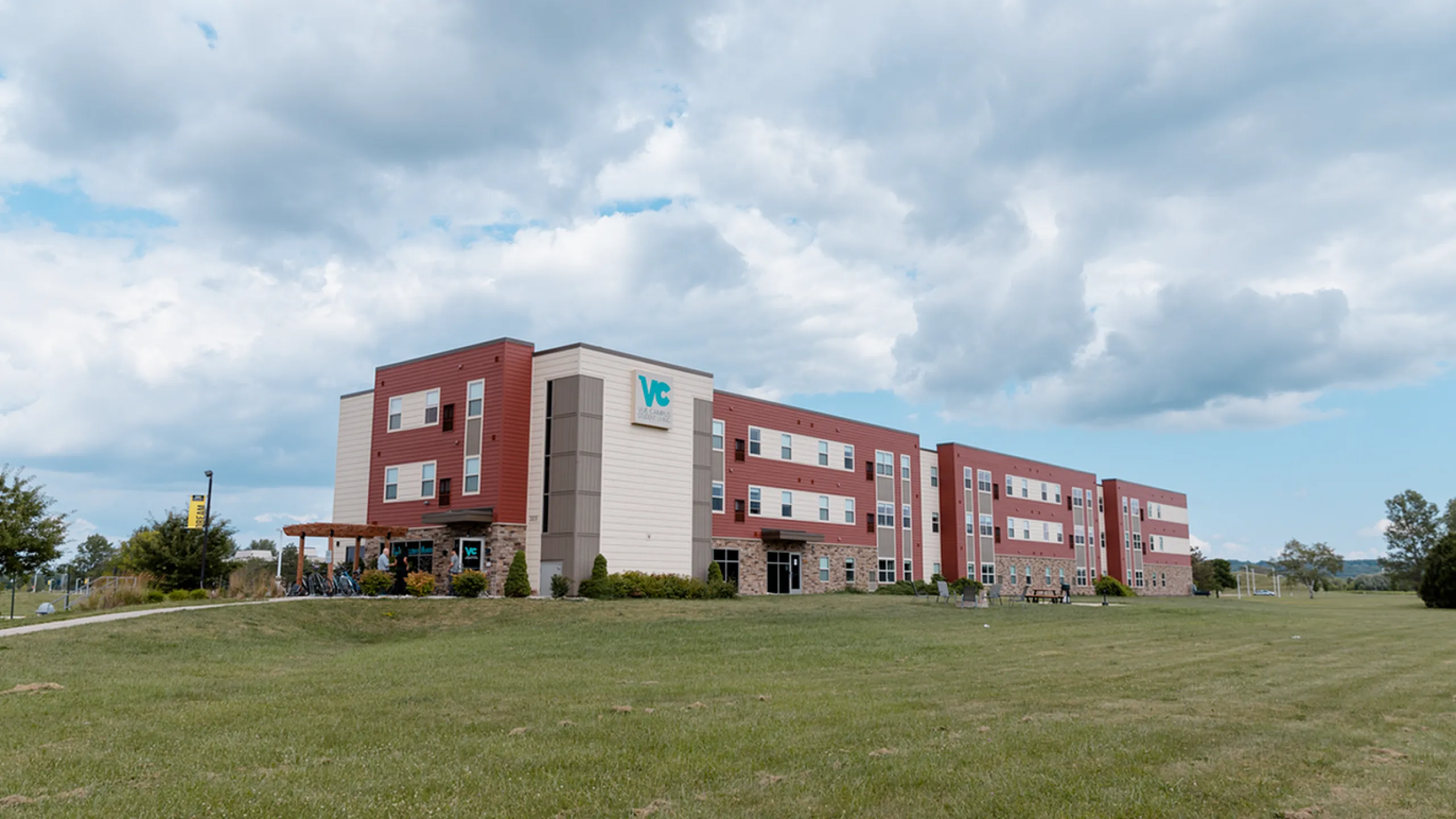 Modern three-story building with red and beige exterior, surrounded by lush green lawn, under a vibrant blue sky.
