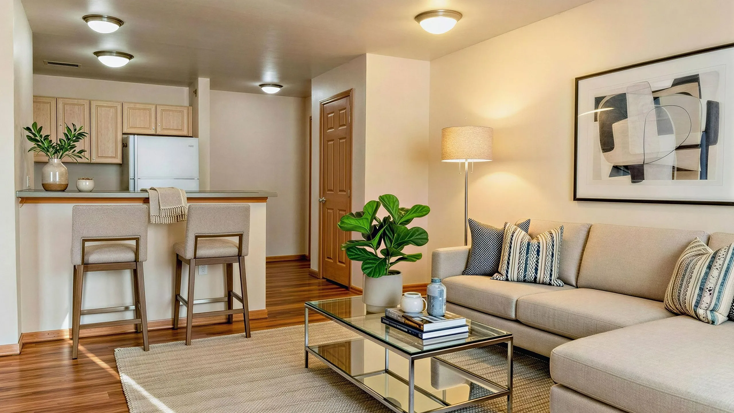 Modern apartment living space featuring a cozy beige sofa, stylish glass coffee table, and inviting kitchen bar with stools, complemented by warm lighting and chic decor.