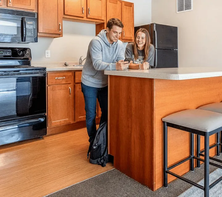 Modern kitchen with warm wooden cabinetry, sleek black appliances, and a cozy breakfast bar. Two people enjoy a bright, inviting space perfect for socializing.
