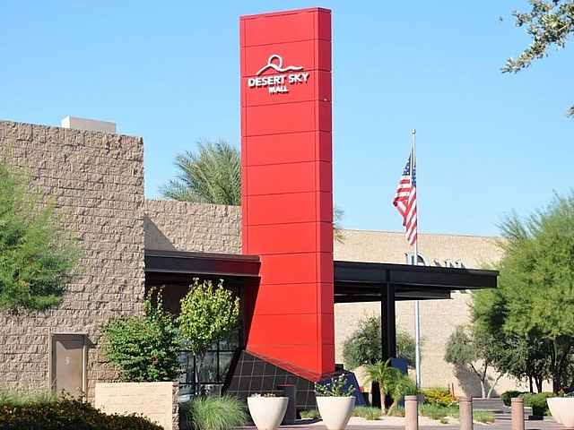 Modern shopping mall entrance featuring bold red architectural design, surrounded by lush greenery and a clear blue sky.