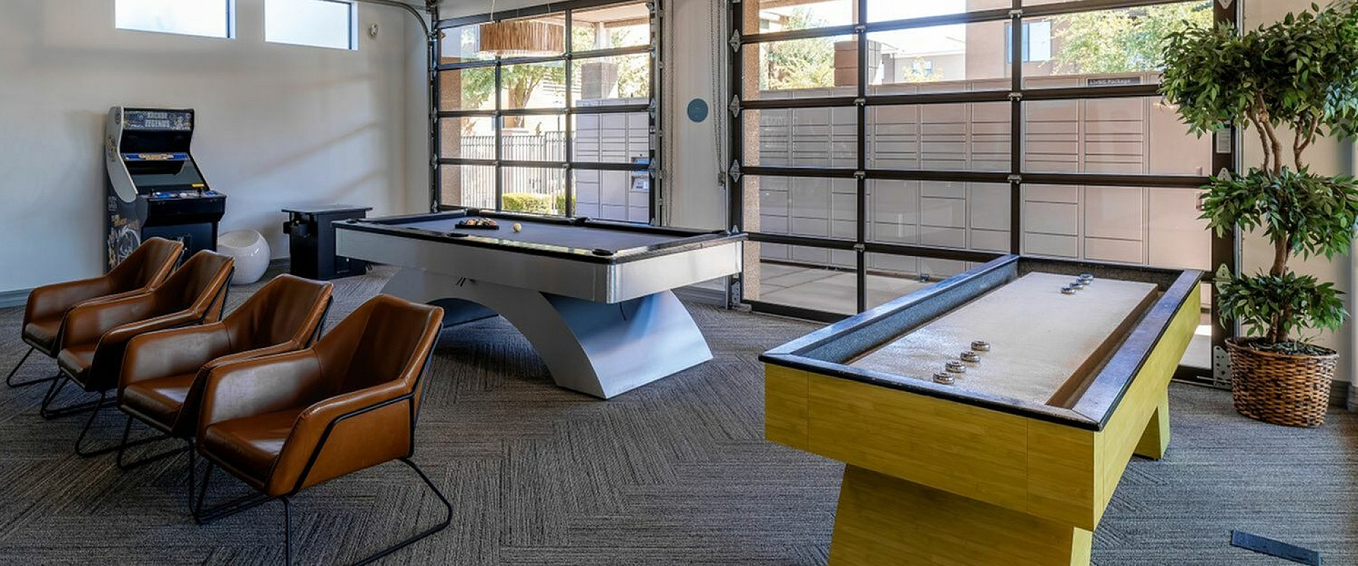 Modern entertainment room featuring luxurious leather chairs, a sleek pool table, vibrant shuffleboard table, and arcade game machine, all set in a bright, open space with large windows.