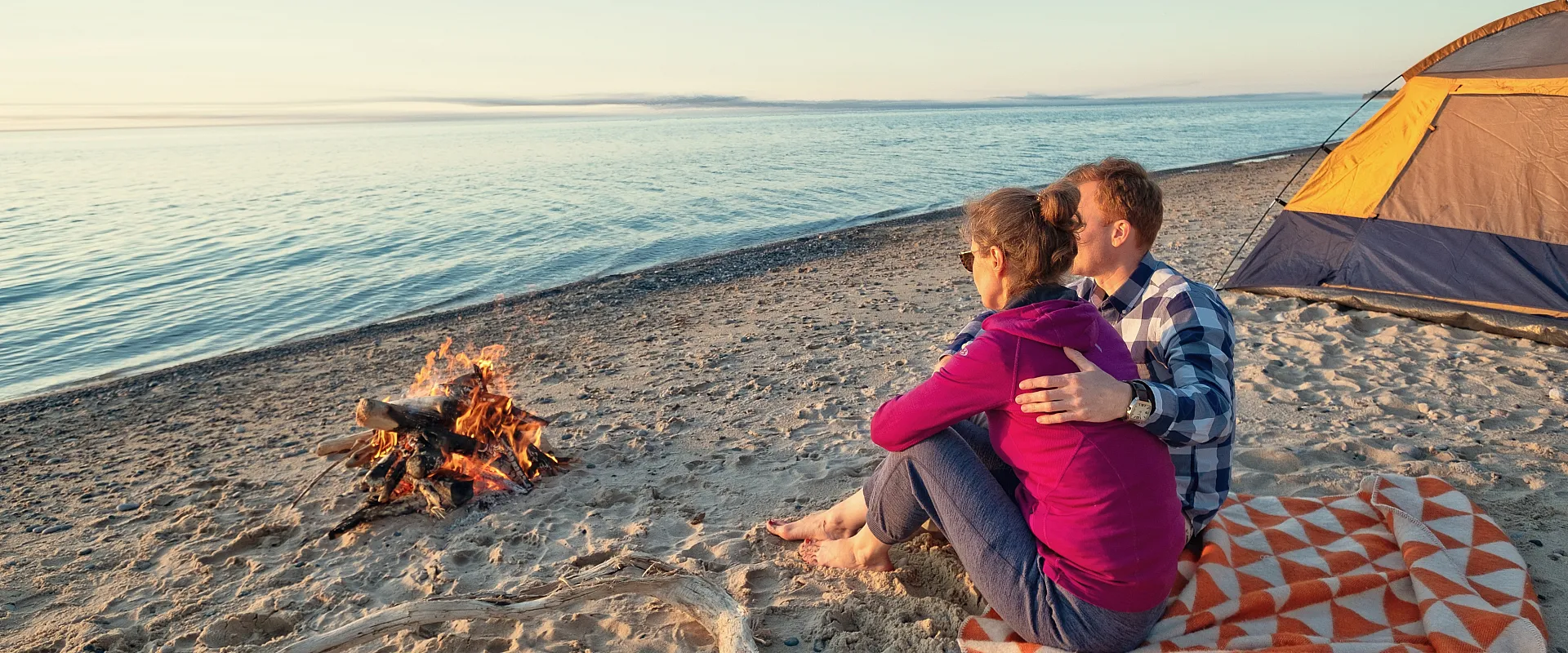 Couple enjoying a serene beach sunset with a cozy campfire and tent, perfect for a peaceful camping getaway.