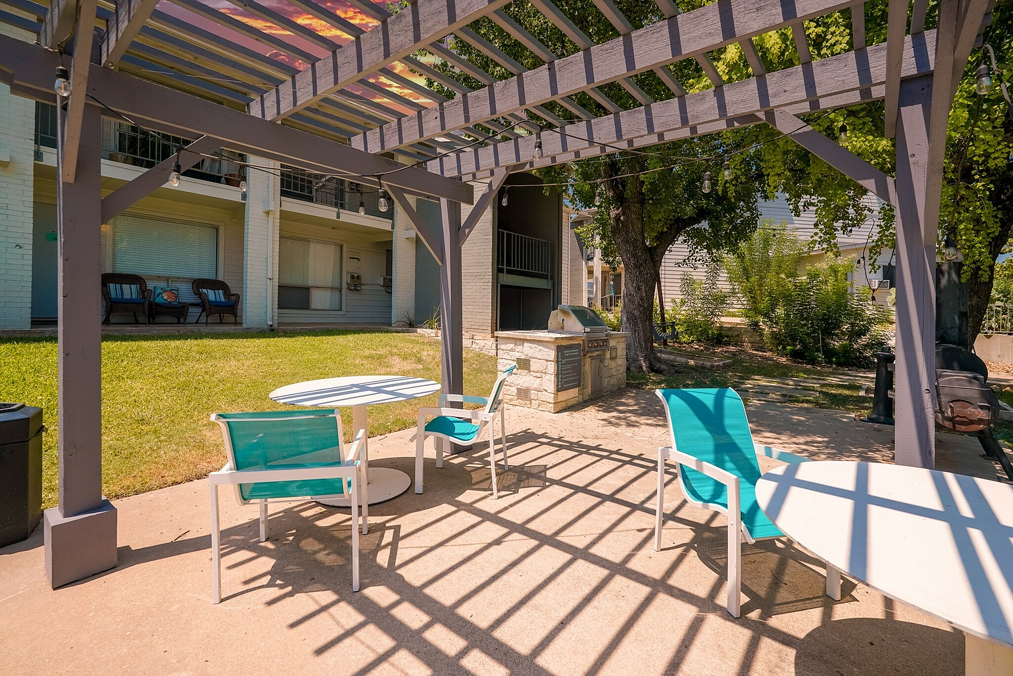 Charming outdoor patio with pergola, colorful seating, and barbecue area amidst lush greenery.