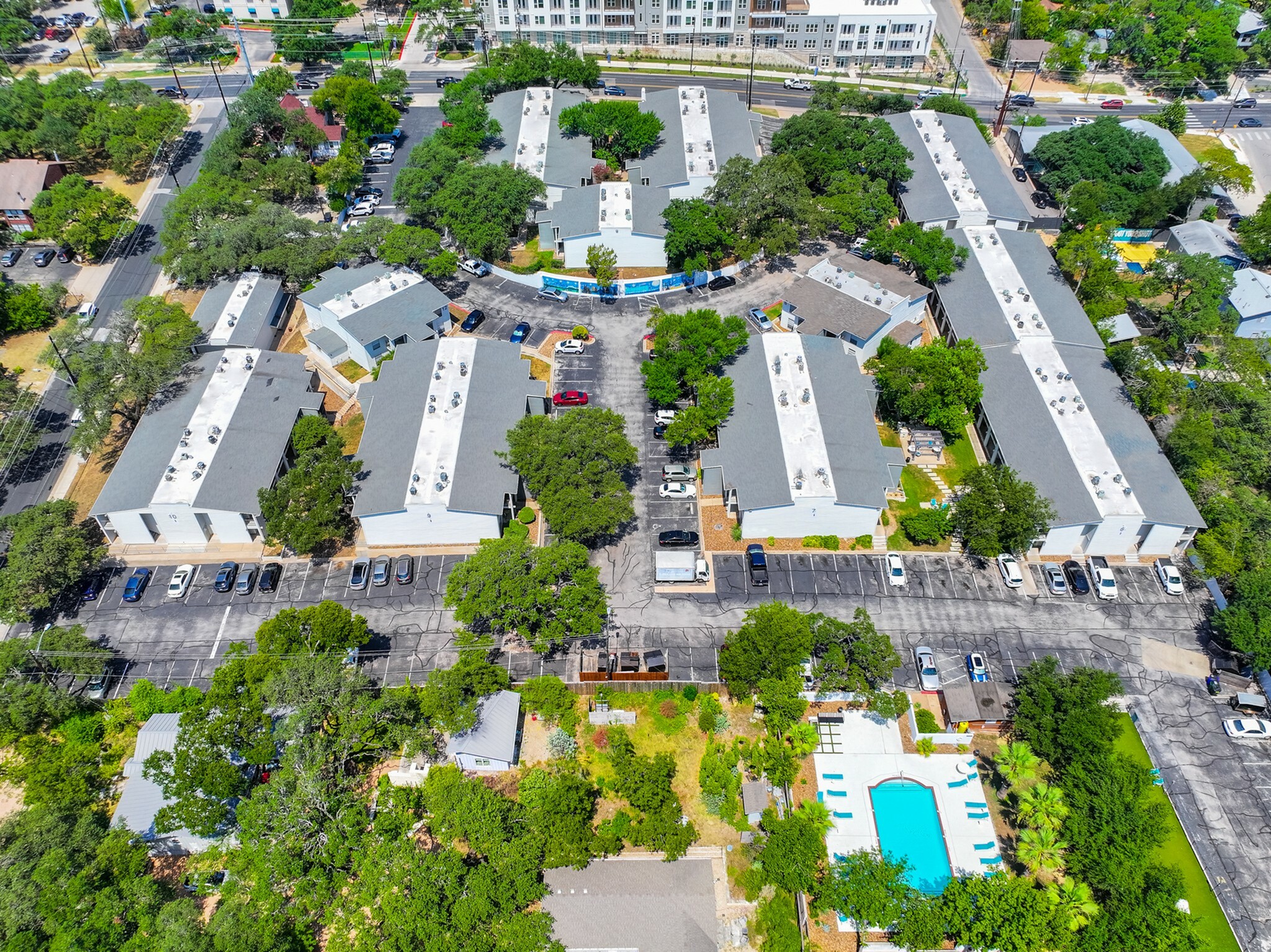 Aerial view of a charming apartment complex with multiple buildings surrounded by lush greenery, ample parking, and inviting swimming pool area.