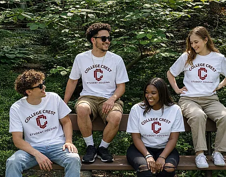 Cheerful group of friends wearing College Crest Student Housing t-shirts, sitting on a bench surrounded by lush greenery.