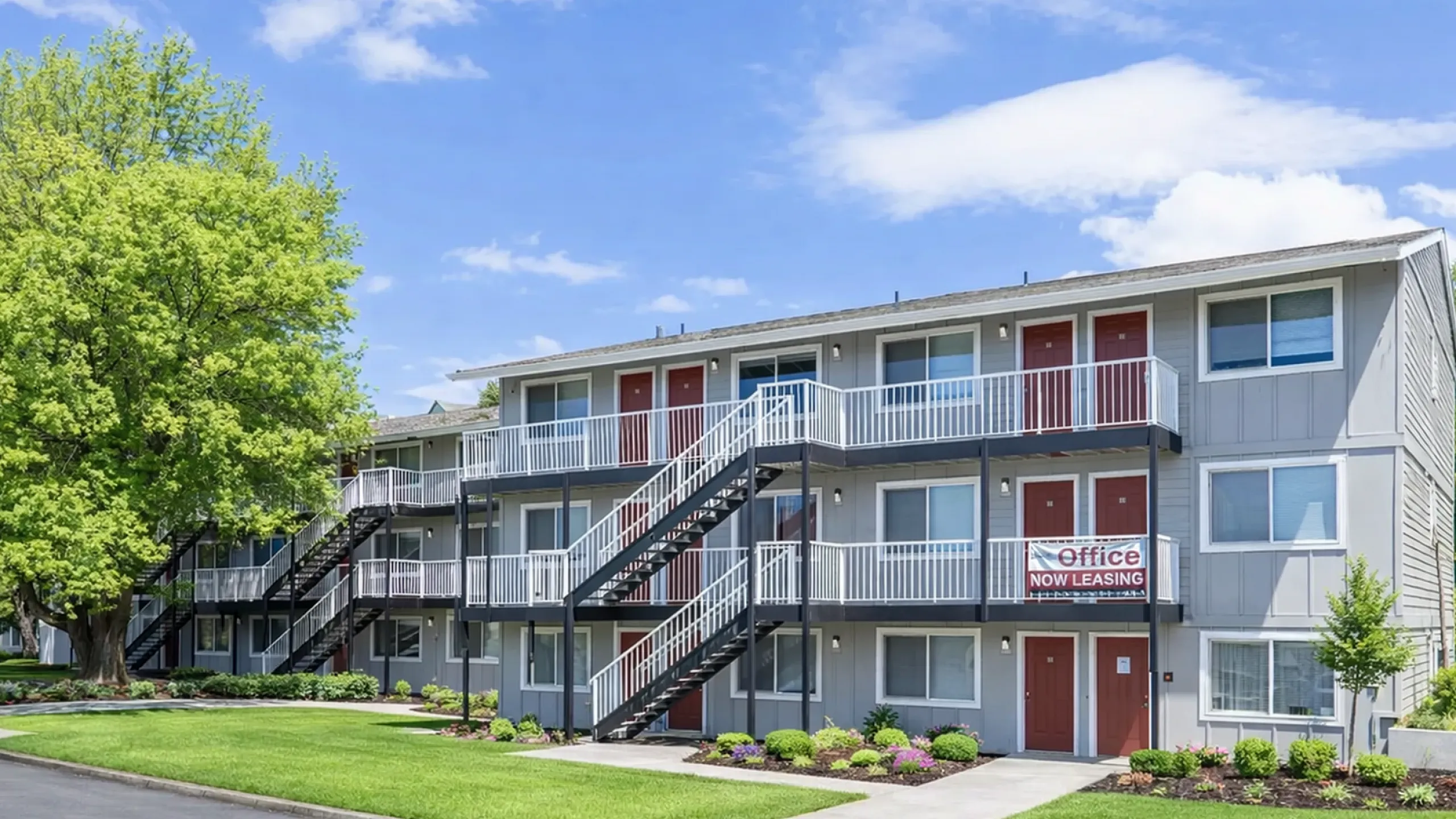 Modern three-story apartment building with red doors, lush landscaping, and a prominent leasing sign.