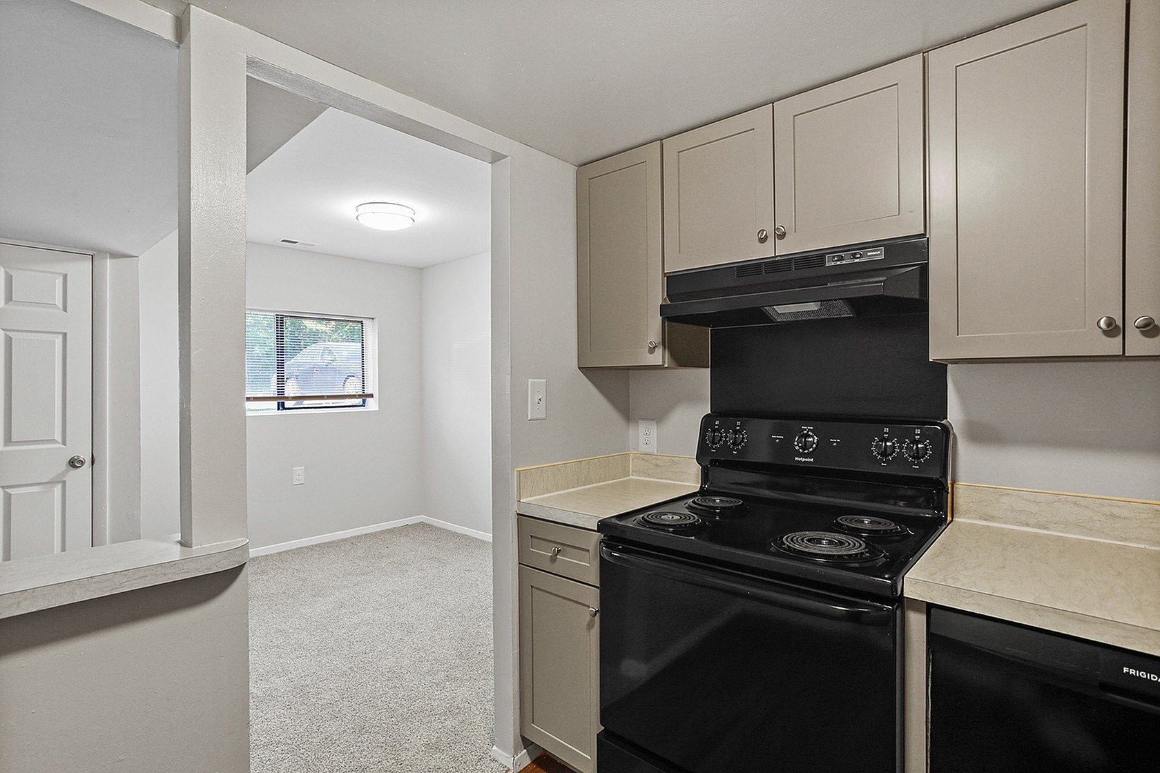 Charming kitchen with sleek beige cabinets, black appliances, and inviting open entry to a sunlit room with a cozy carpeted floor.