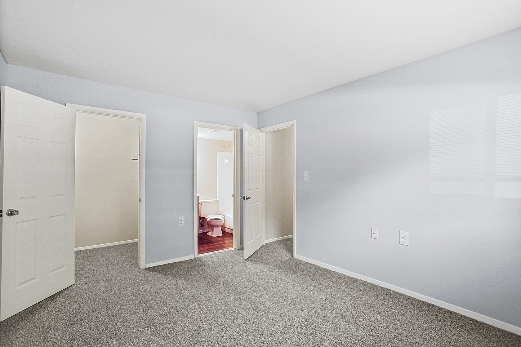 Bright and spacious bedroom featuring neutral walls, plush carpet, and a glimpse of an en-suite bathroom through an open door.