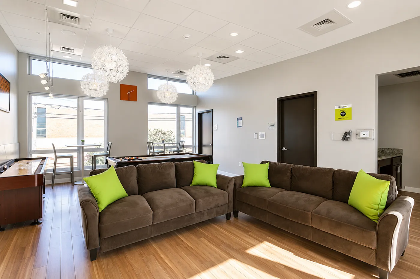 Bright and inviting lounge area featuring modern decor, cozy brown sofas with vibrant green pillows, elegant lighting fixtures, and large windows offering plenty of natural light.