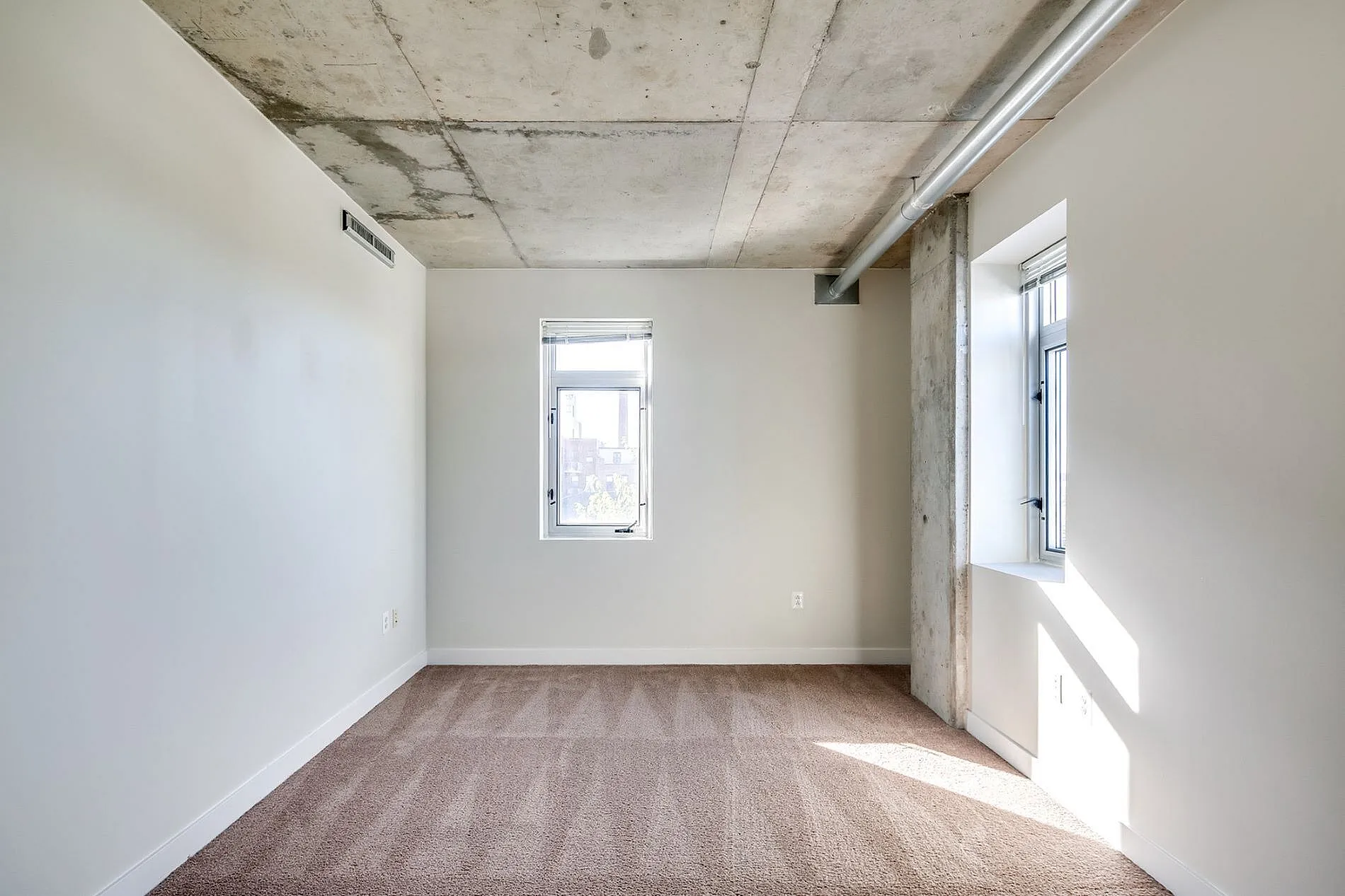 Bright and airy room with modern concrete ceiling, large windows, and plush carpeting, perfect for a cozy retreat.