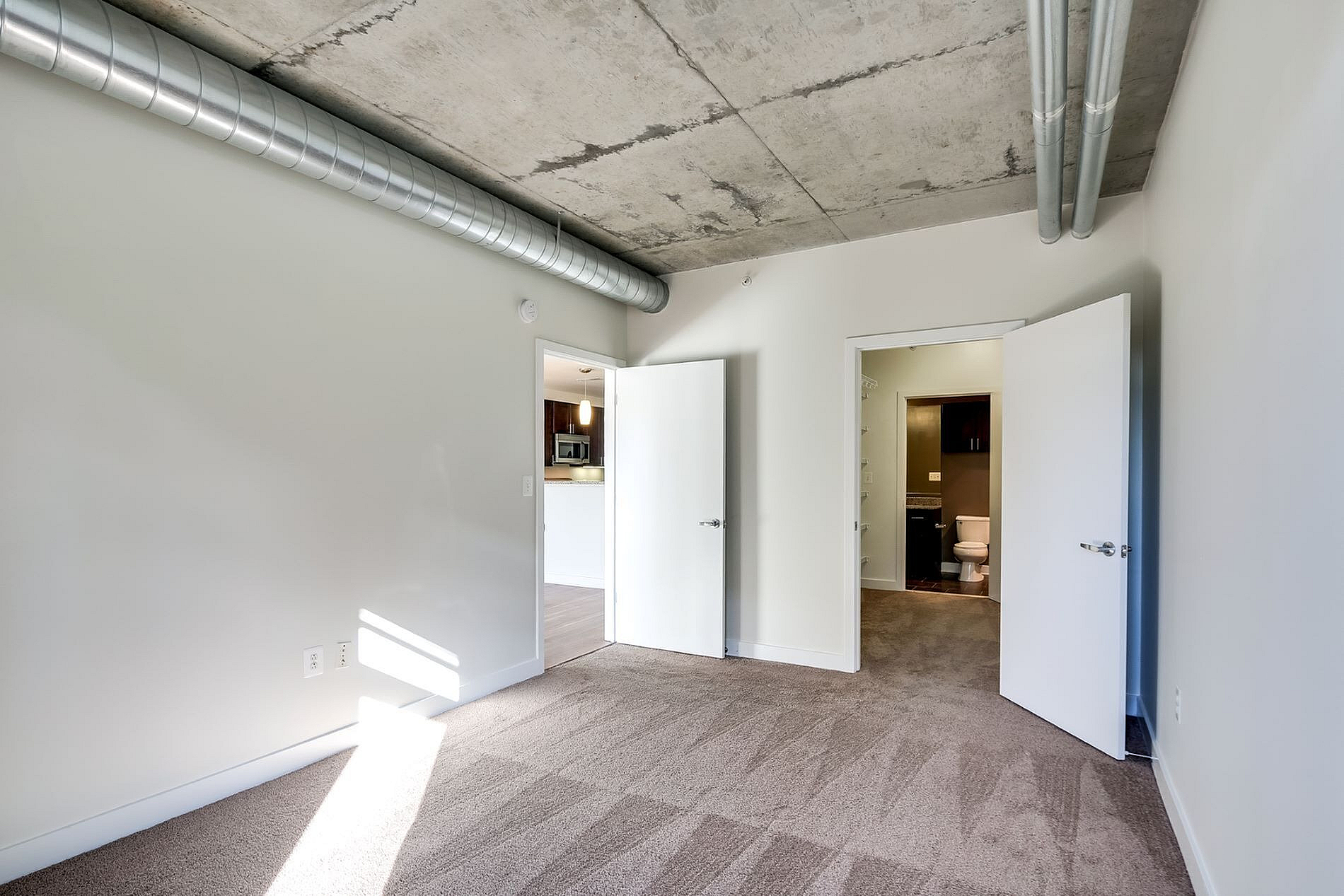 Contemporary apartment featuring a spacious room with stylish exposed ductwork, plush carpeting, and ample natural light, leading to a modern kitchen and bathroom.