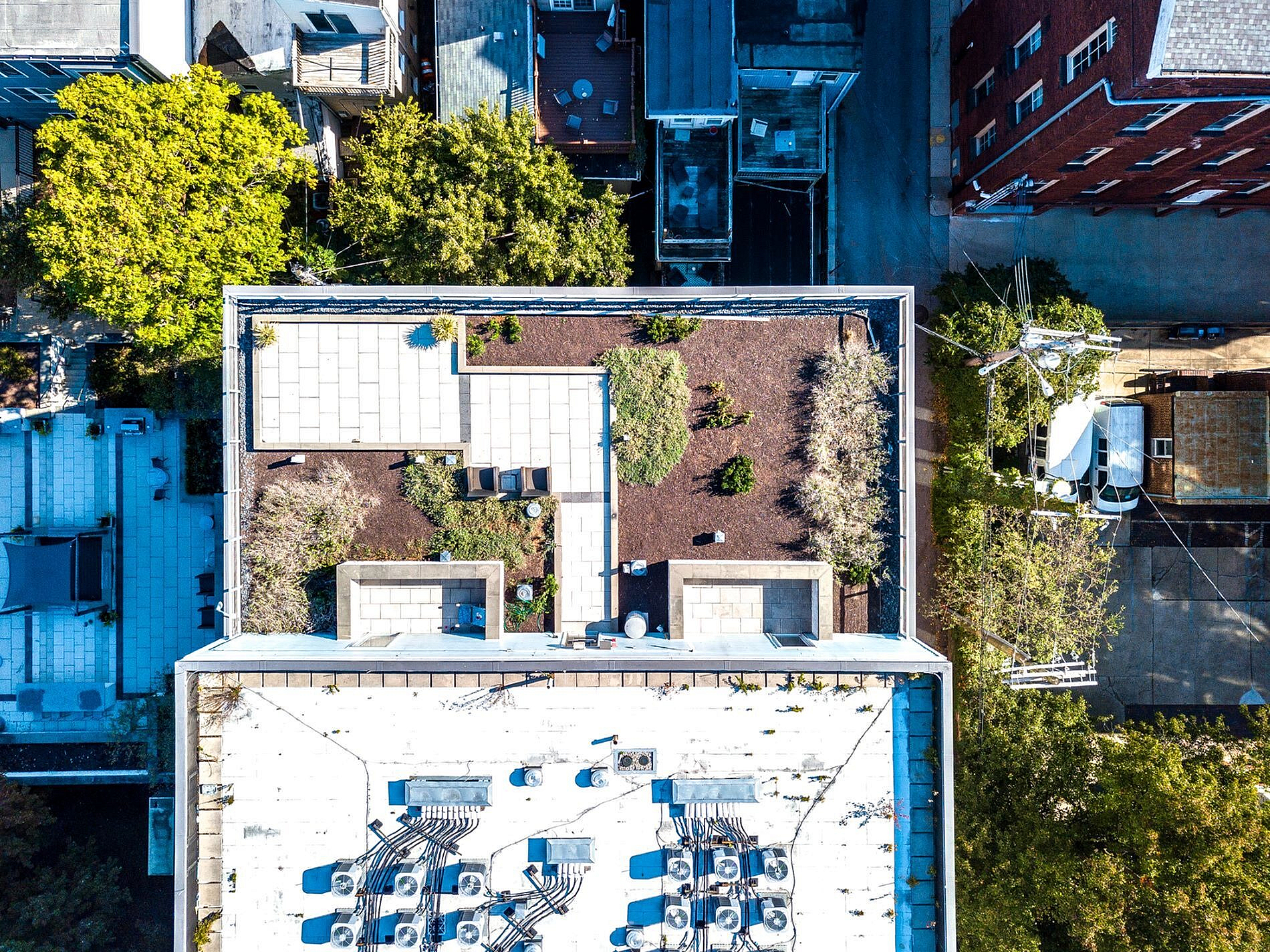 Aerial view of a modern rooftop garden with lush greenery and elegant seating area, surrounded by urban architecture and vibrant trees.