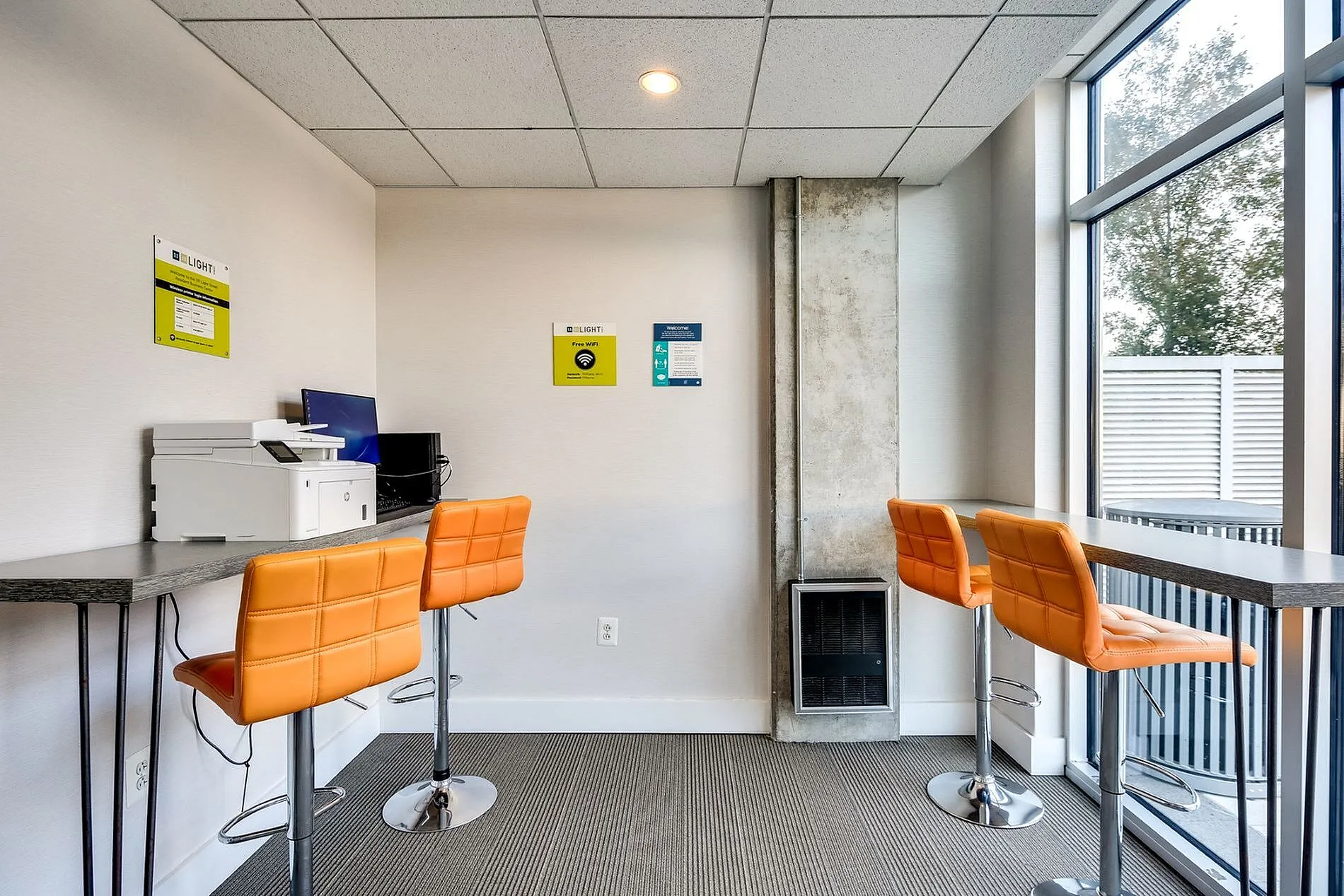 Modern apartment business center with vibrant orange bar stools, sleek workstations, and large windows for natural light.