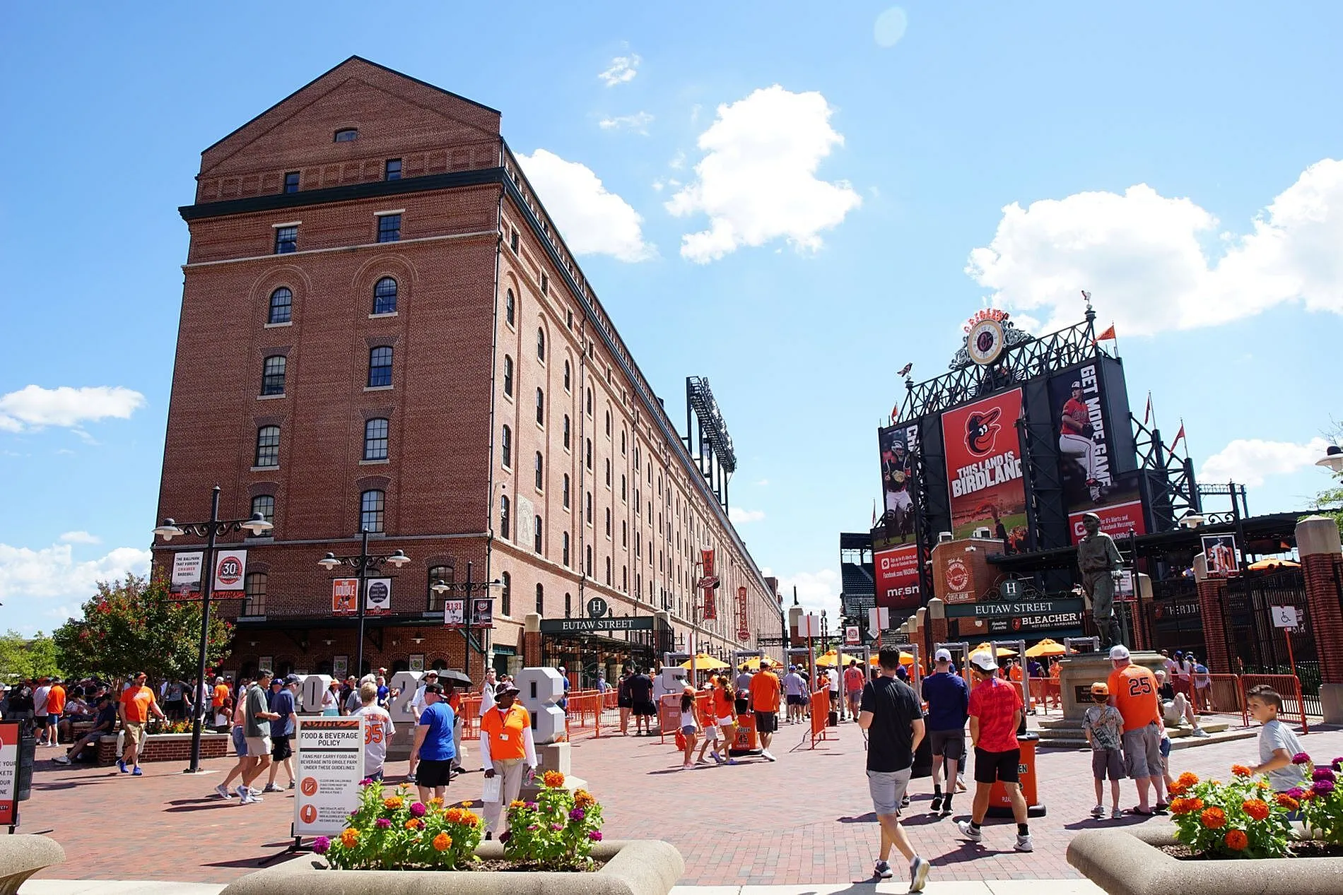 Charming urban scene with a historic red-brick building, vibrant crowds enjoying a sunny day, and a welcoming plaza with colorful flowers.