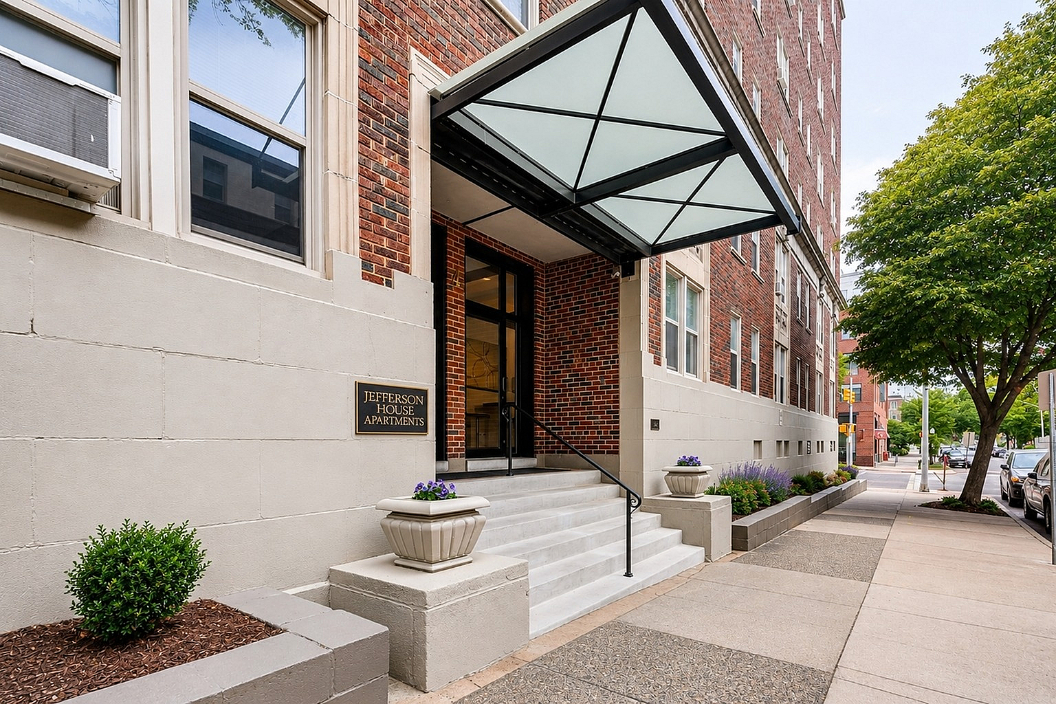 Inviting entrance to Jefferson House Apartments with elegant modern canopy, lush greenery, and charming brick facade, offering a glimpse of urban living at its finest.