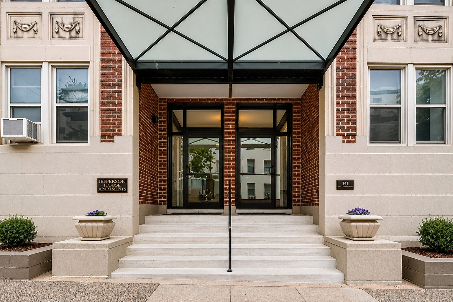 Elegant entrance of Jefferson House Apartments, featuring modern glass doors, red brick facade, and stylish planters.