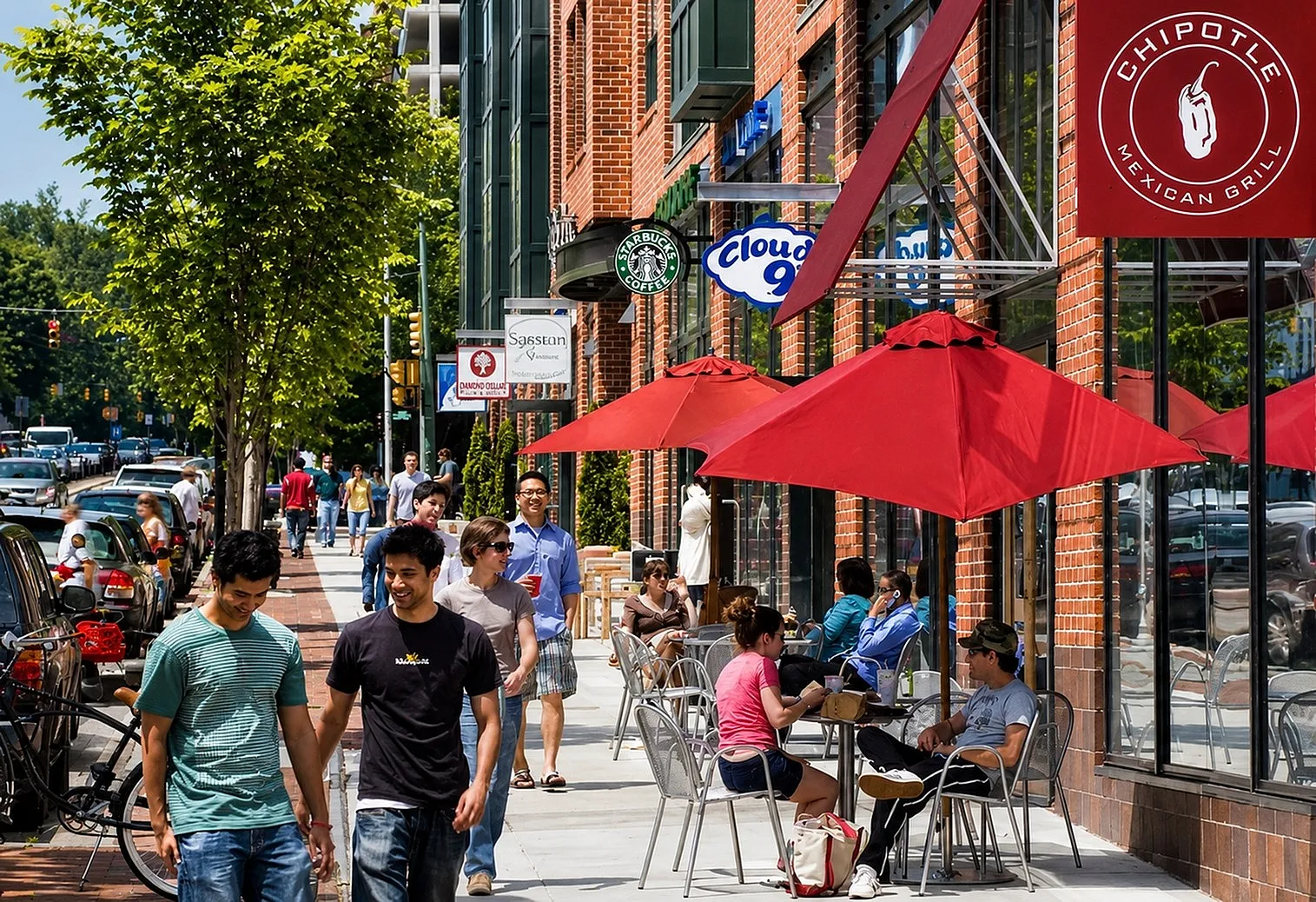Vibrant urban street scene with people enjoying a sunny day at outdoor cafes under bright red umbrellas. Charming brick storefronts and lush green trees line the bustling sidewalk.