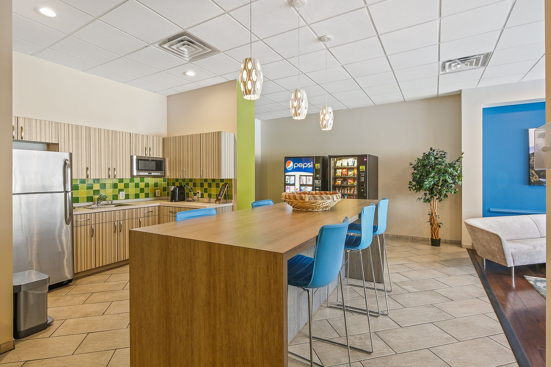 Modern community kitchen with stylish green tile backsplash, sleek stainless steel appliances, and a spacious island with vibrant blue seating, offering a welcoming atmosphere.