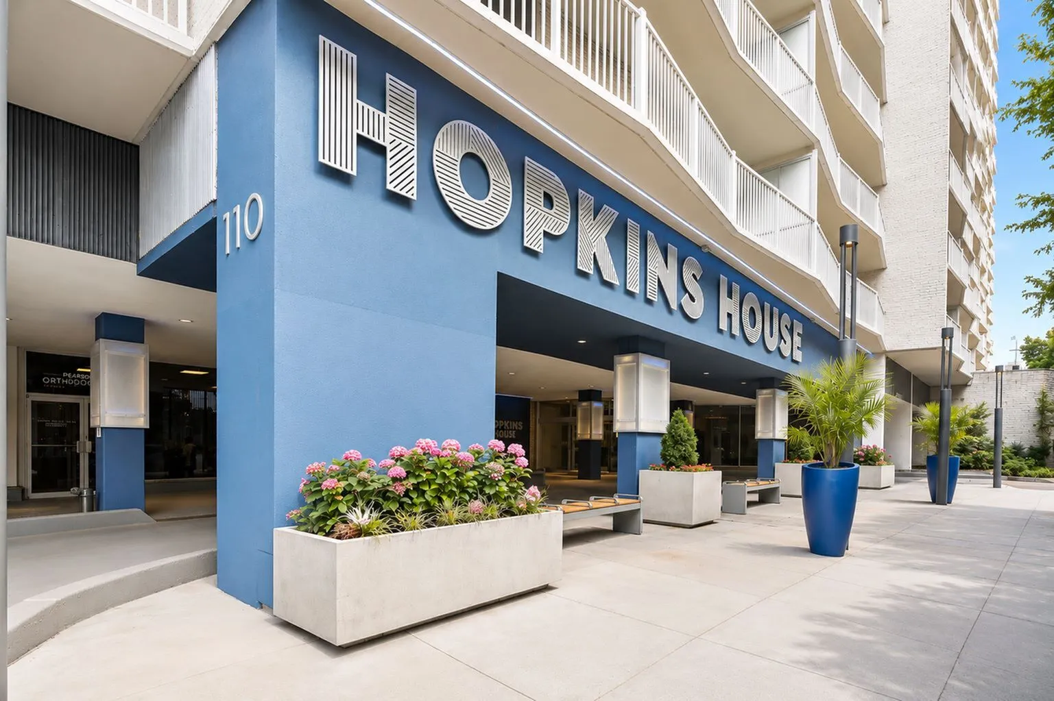 Modern high-rise exterior of Hopkins House, featuring sleek blue facade and elegant landscaping with vibrant potted plants.