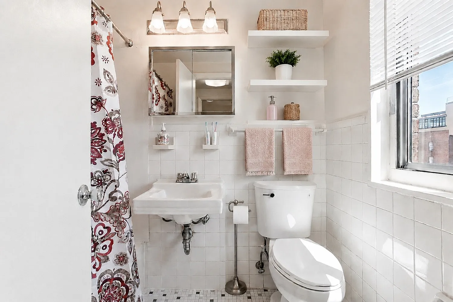 Charming bathroom featuring elegant lighting, floral shower curtain, and crisp white tiles with modern shelving.
