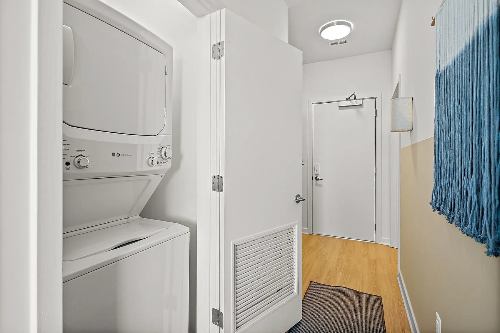Bright and modern apartment entryway featuring an in-unit washer and dryer, wooden flooring, and chic textile wall art.
