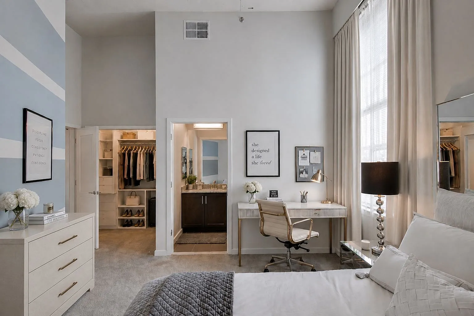 Elegant, spacious bedroom with high ceilings, featuring a cozy workspace, ample natural light, and a walk-in closet leading to an en-suite bathroom.