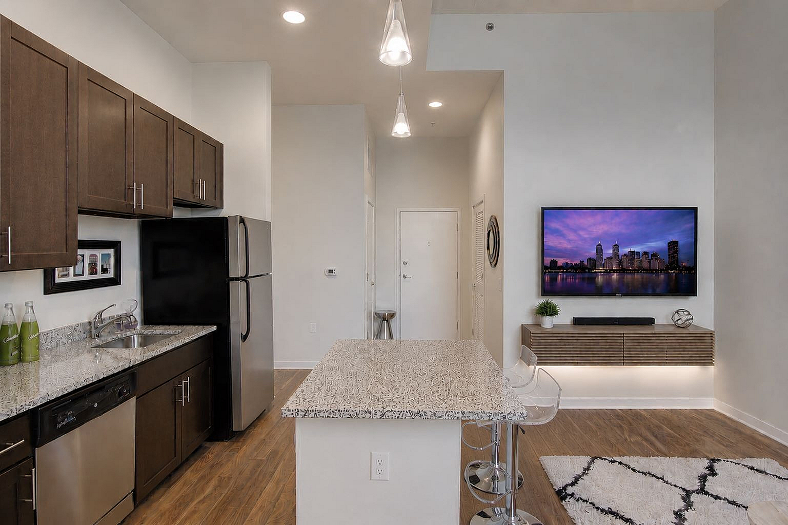 Contemporary kitchen and living area featuring sleek brown cabinets, stainless steel appliances, stylish island counter, and wall-mounted TV with city skyline view.