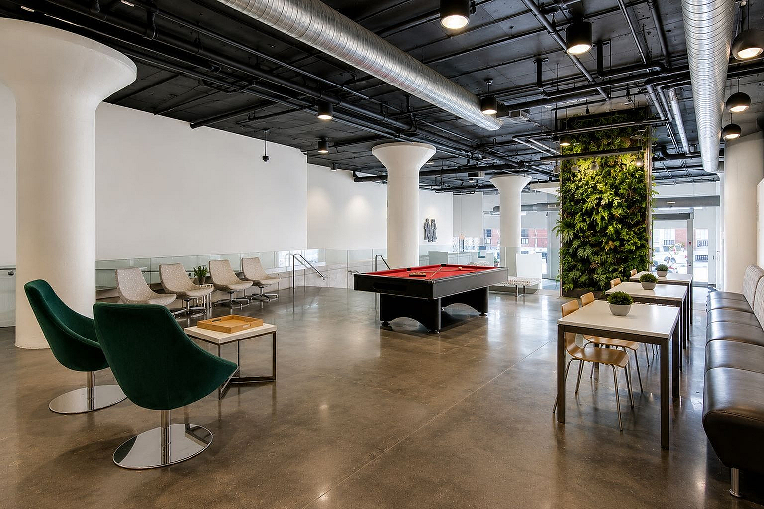 Modern communal space boasting sleek seating, a vibrant green wall, and a stylish billiards table under industrial-chic ceilings.