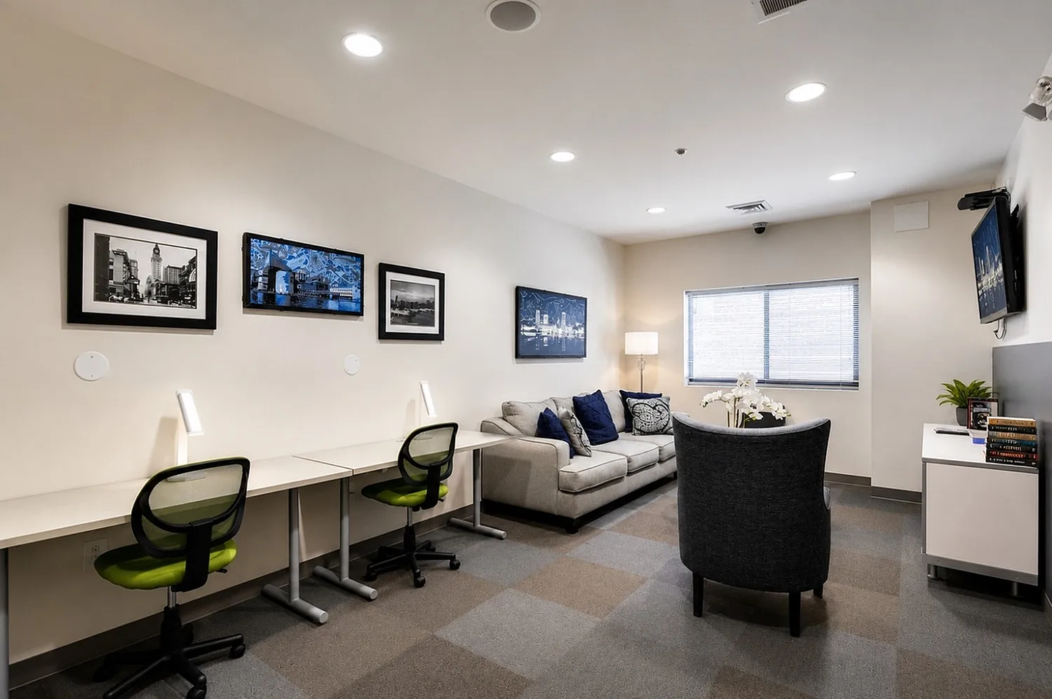Modern lounge area with stylish decor, featuring a cozy sofa, artistic wall frames, and a functional workspace with green chairs.