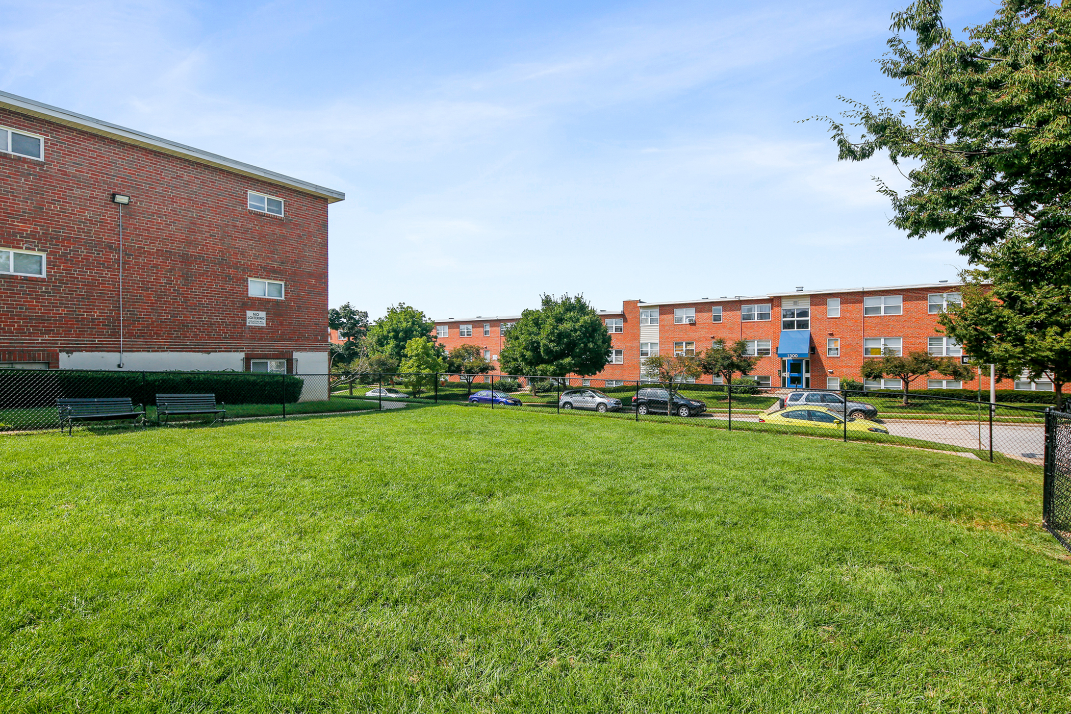 Charming apartment complex with lush green lawn, inviting benches, and red brick buildings under a clear blue sky.