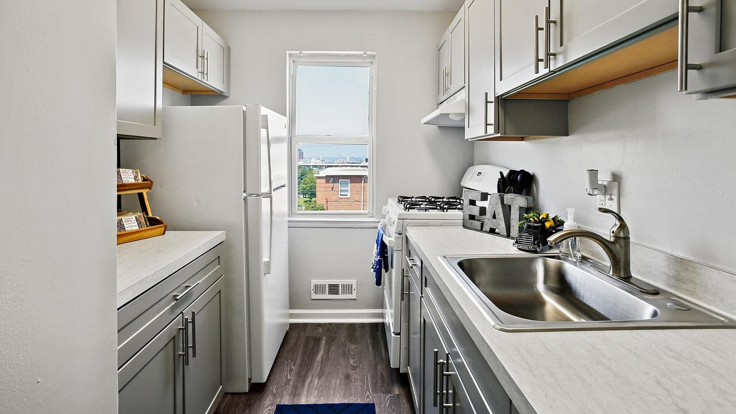 Bright and modern galley kitchen with sleek white cabinets, stainless steel sink, and a large window offering city views.