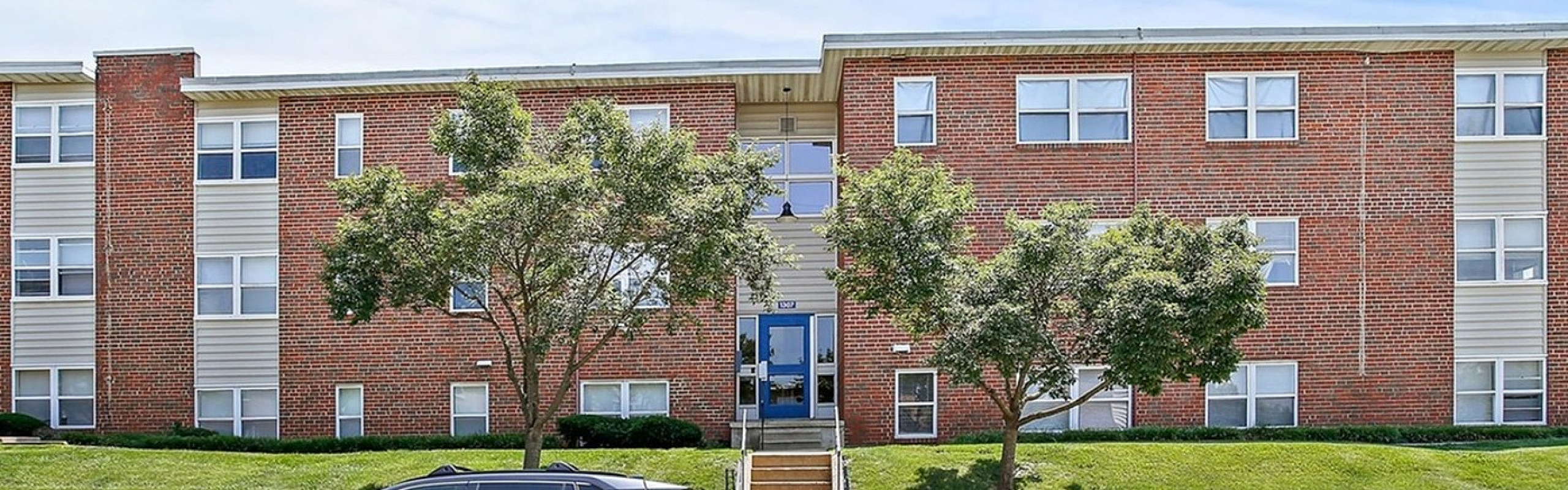 Charming three-story brick apartment building with lush green lawns and mature trees, perfect for serene suburban living.