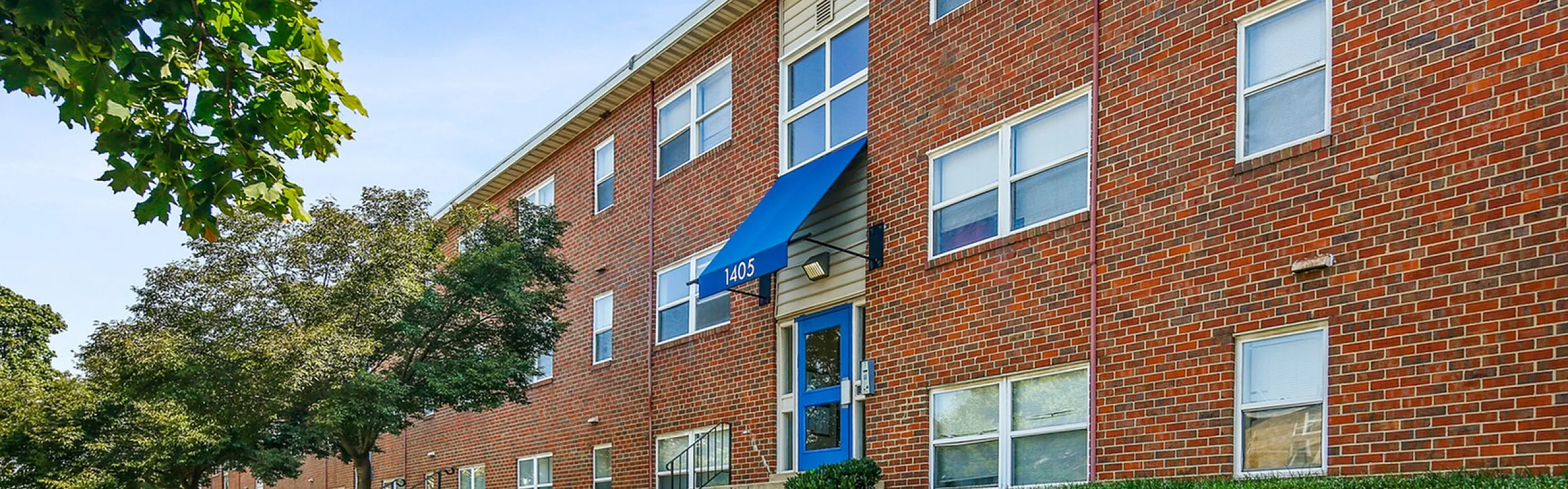 Charming brick apartment building with lush green landscaping and a vibrant blue awning entrance.