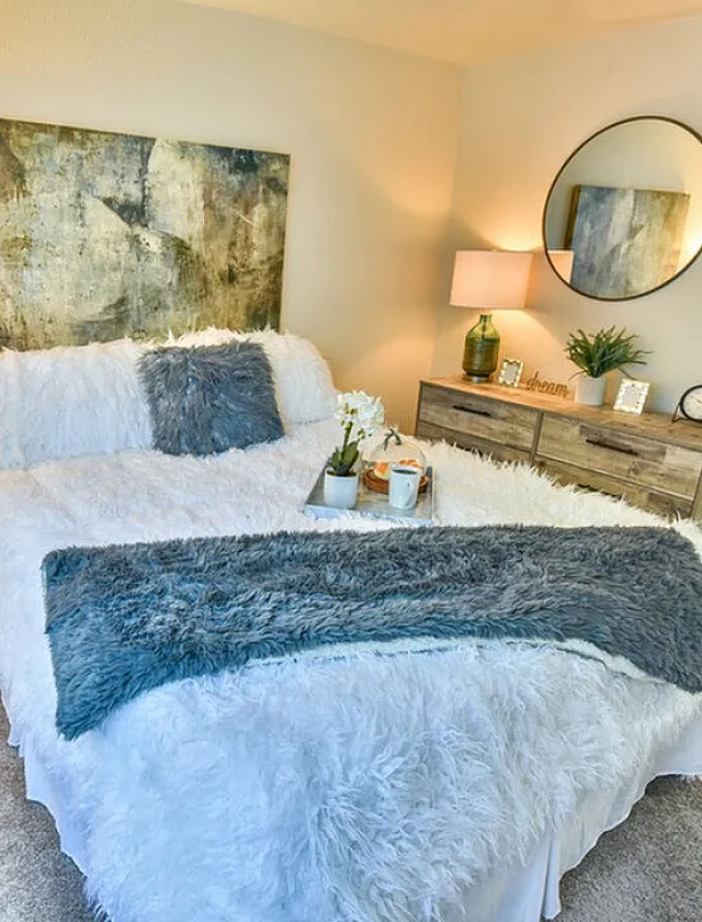 Cozy bedroom with elegant decor, featuring a plush bed with fluffy blankets, stylish side lamps, and modern art on light-colored walls.