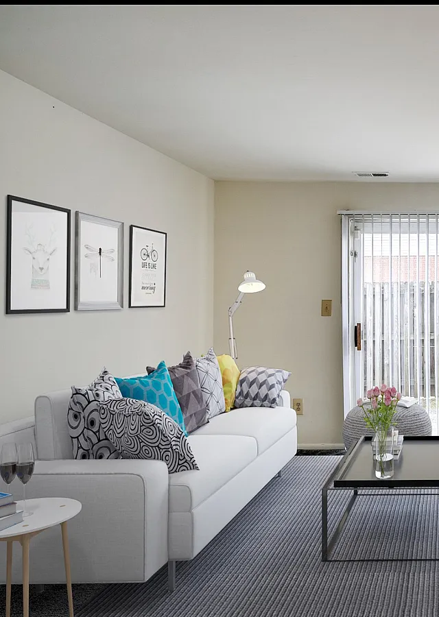 Bright and airy living room with modern decor, white sofa with colorful cushions, sleek coffee table, and natural light streaming through vertical blinds.