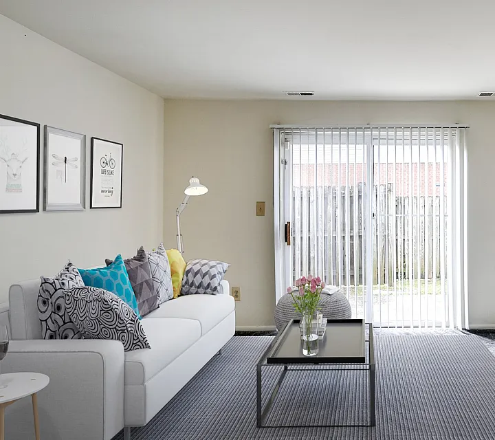 Modern living room with a cozy white sofa adorned with colorful pillows, contemporary wall art, and a sleek TV unit. Sunlit space with a large sliding glass door leading to a private outdoor area, enhanced by stylish decor and a plush rug.