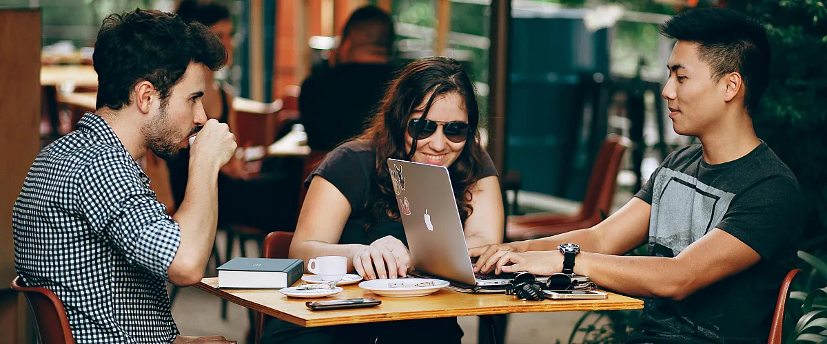 Vibrant outdoor cafe setting with three friends enjoying conversation and shared work on a laptop, surrounded by lush greenery.