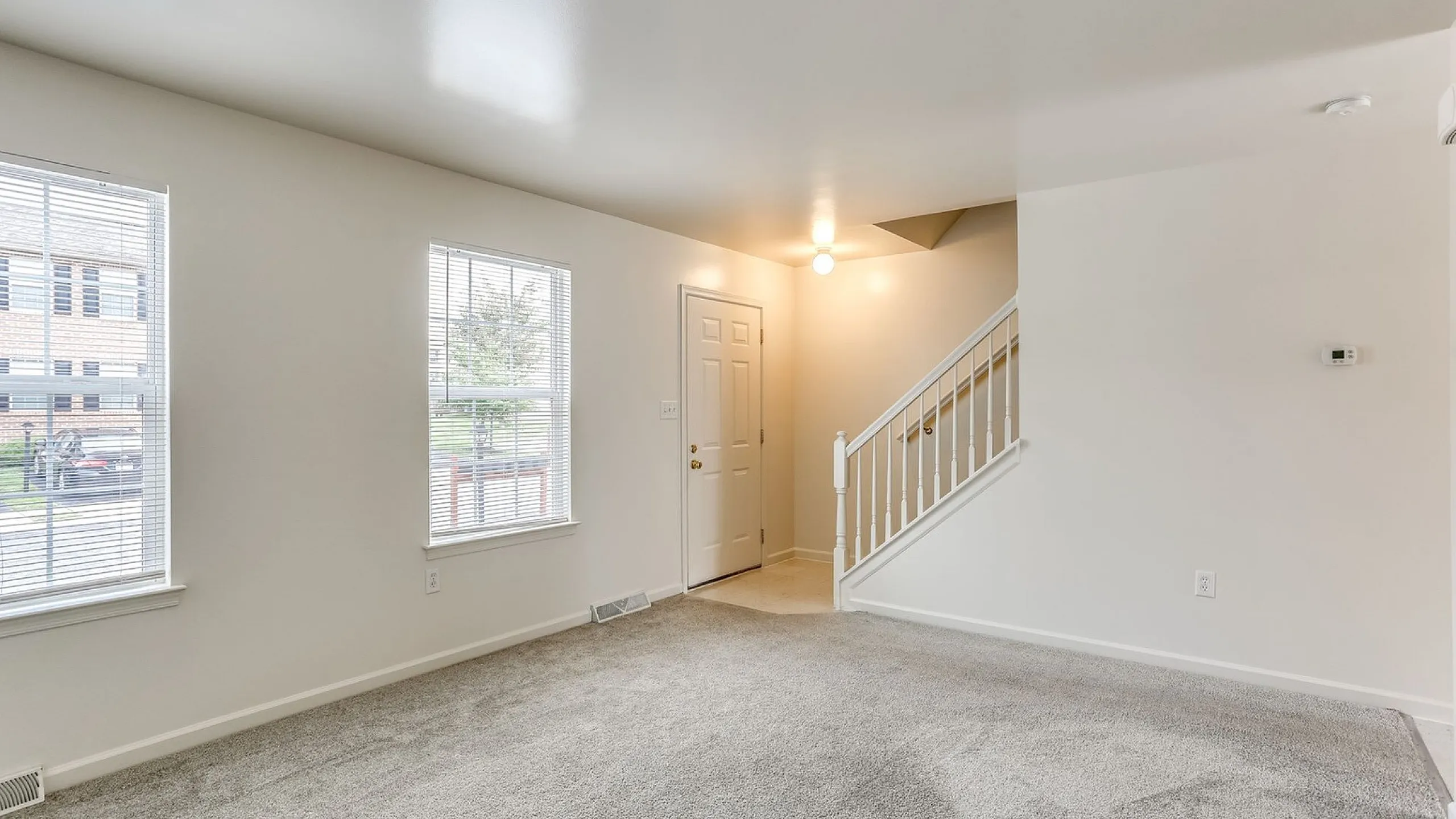 Bright living area featuring soft carpet flooring and natural light from large windows, with a cozy staircase leading to the upper floor.