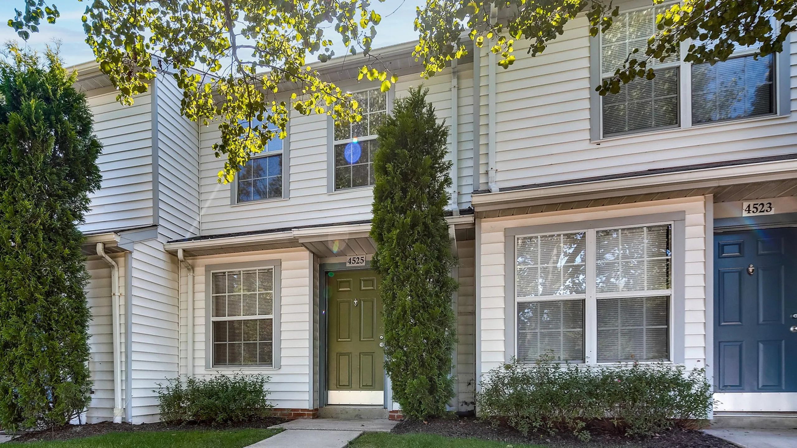 Charming townhouse with lush green entrance, stylish white facade, and inviting front door under a canopy of trees.
