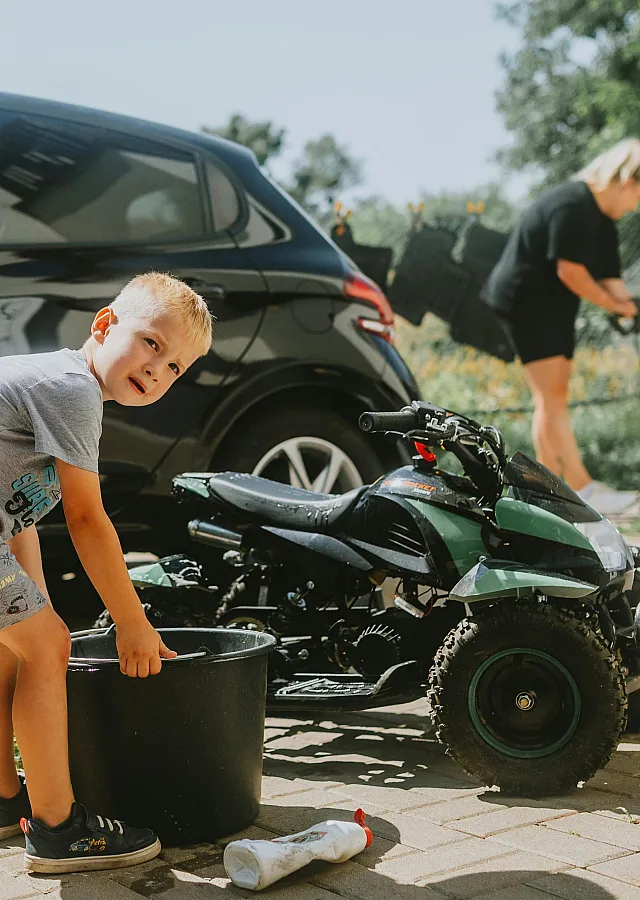A sunny day car cleaning scene with a child assisting playfully near a sleek black car and an ATV.