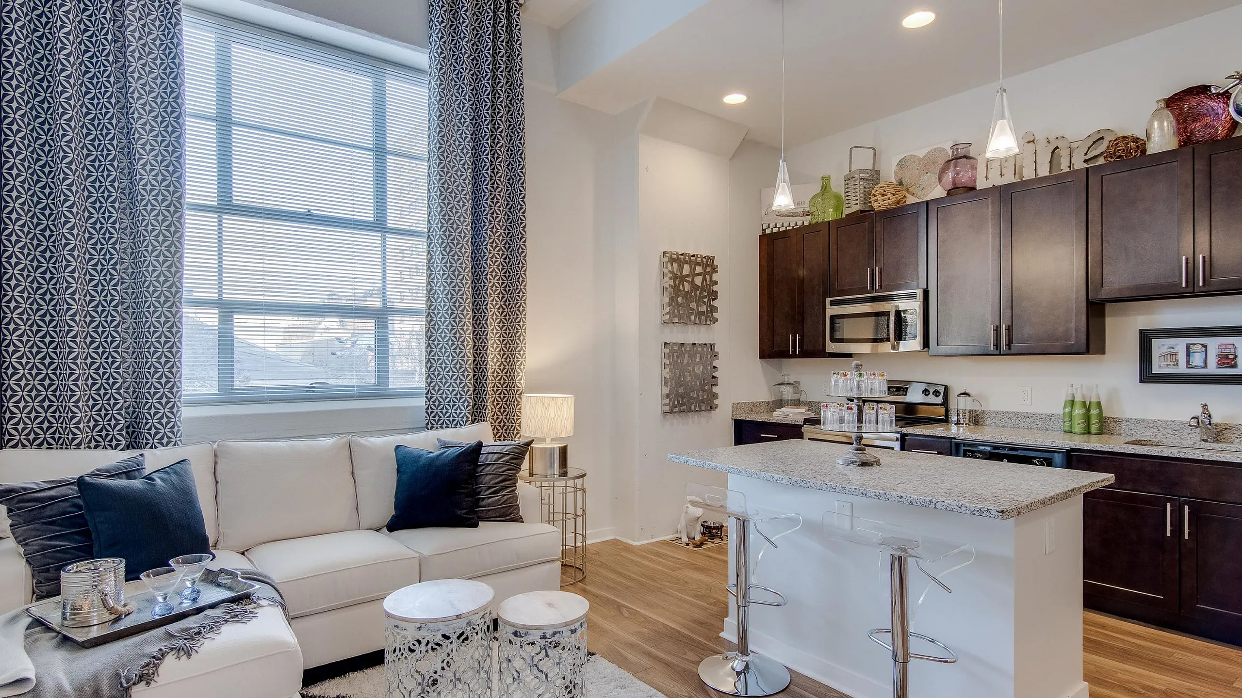 Modern open-concept living room and kitchen with elegant d&eacute;cor, featuring a cozy white sofa, stylish bar stools, and chic dark wood cabinetry.