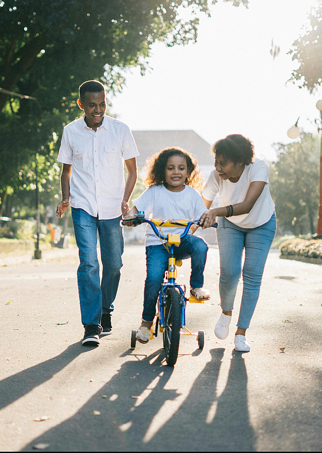 Joyful family teaching a child to ride a bicycle on a sunny day, showcasing an ideal neighborhood for family recreation.