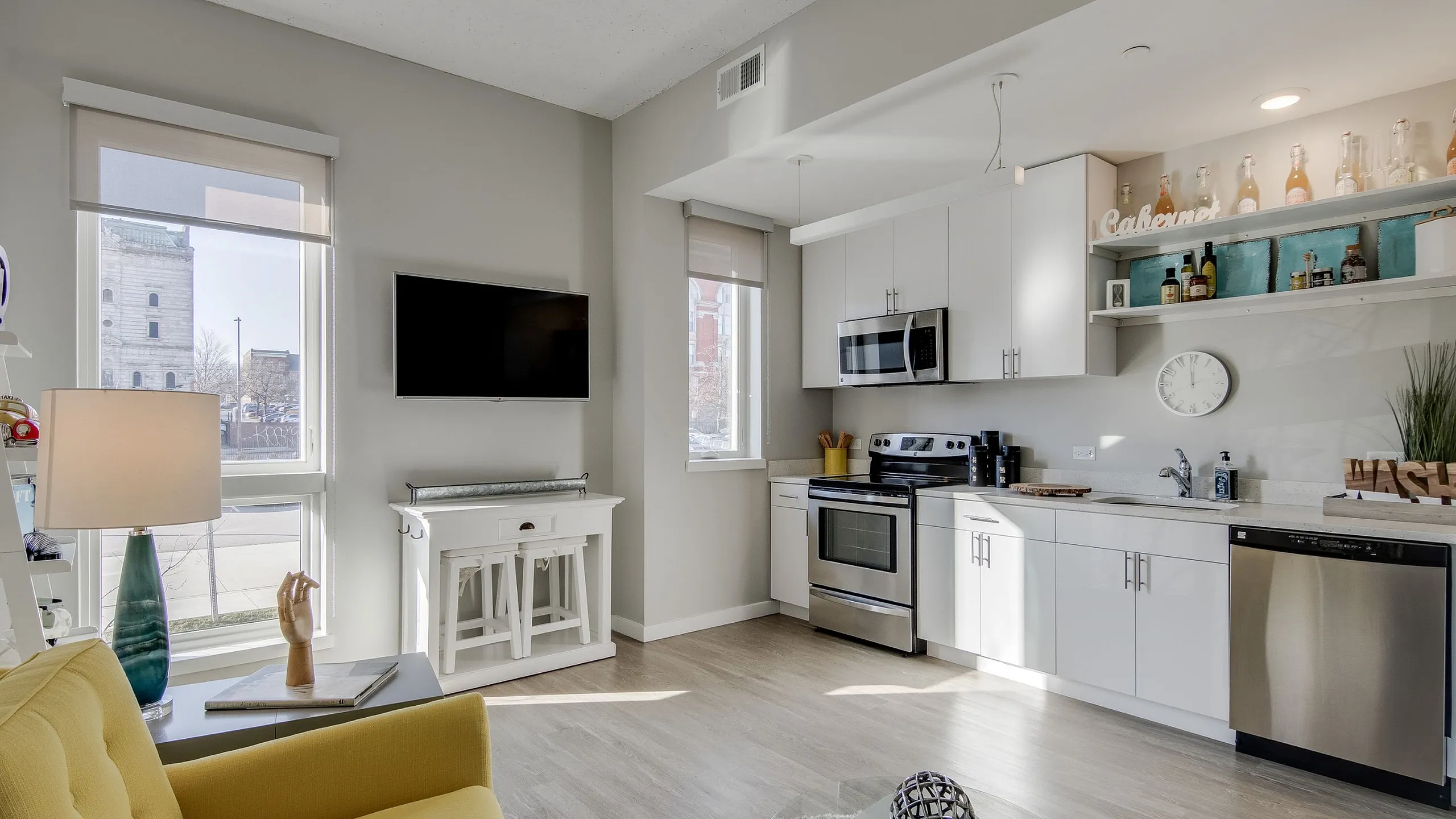 Bright and modern living space featuring an open kitchen with sleek white cabinetry, stainless steel appliances, and a cozy sitting area with a stylish yellow chair, complemented by abundant natural light and contemporary decor.