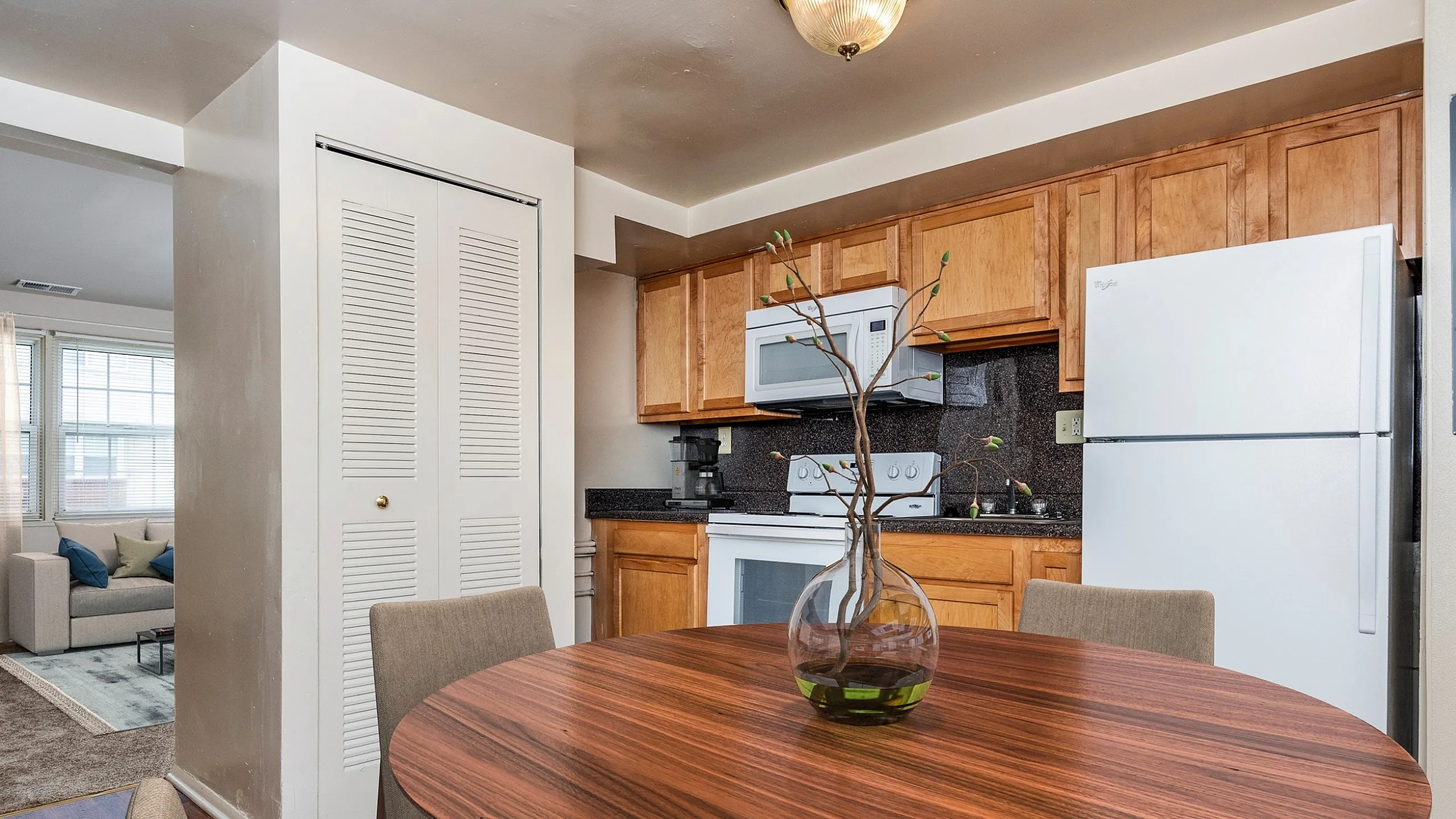 Charming apartment dining area with a round wooden table leading into a cozy, well-lit living space and modern kitchen with wooden cabinetry.