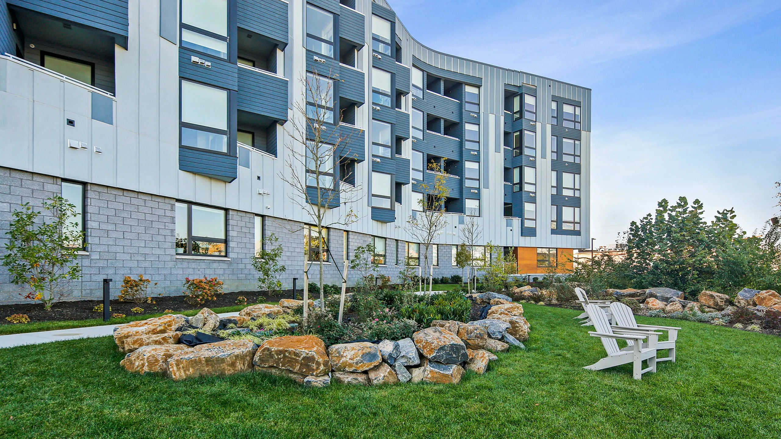 Modern apartment building with stylish architecture, featuring extensive landscaped grounds with seating areas and lush greenery.