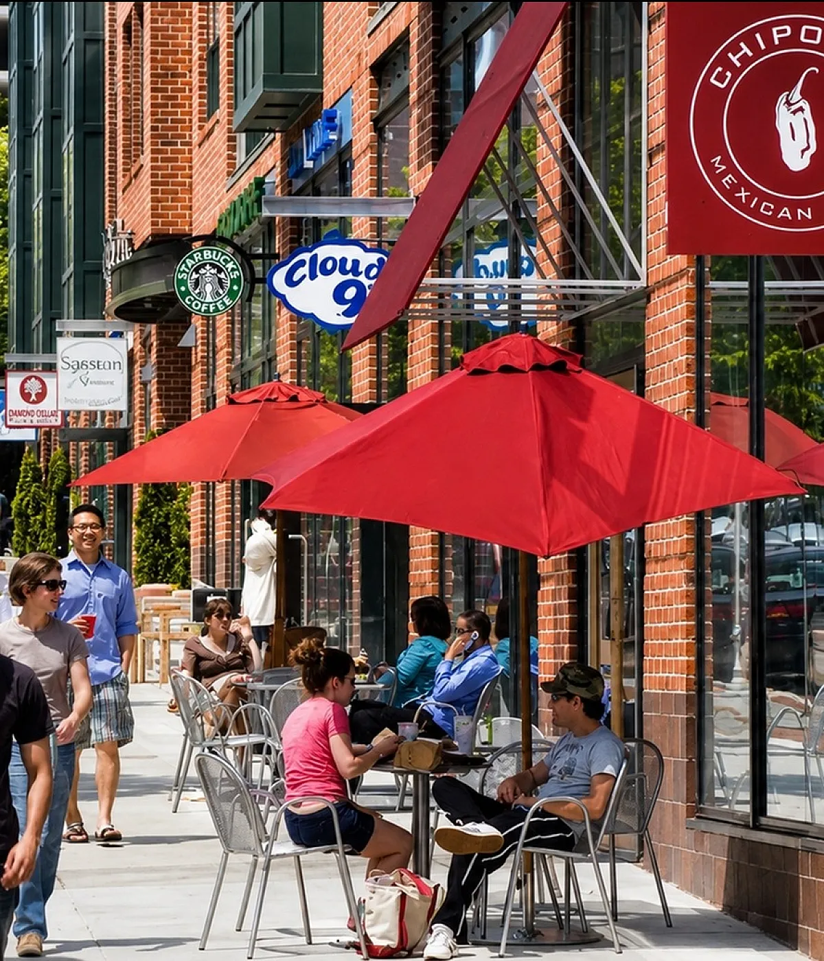 Vibrant urban setting with bustling sidewalk cafes, red umbrellas, and pedestrians enjoying a lively atmosphere outside popular shops.
